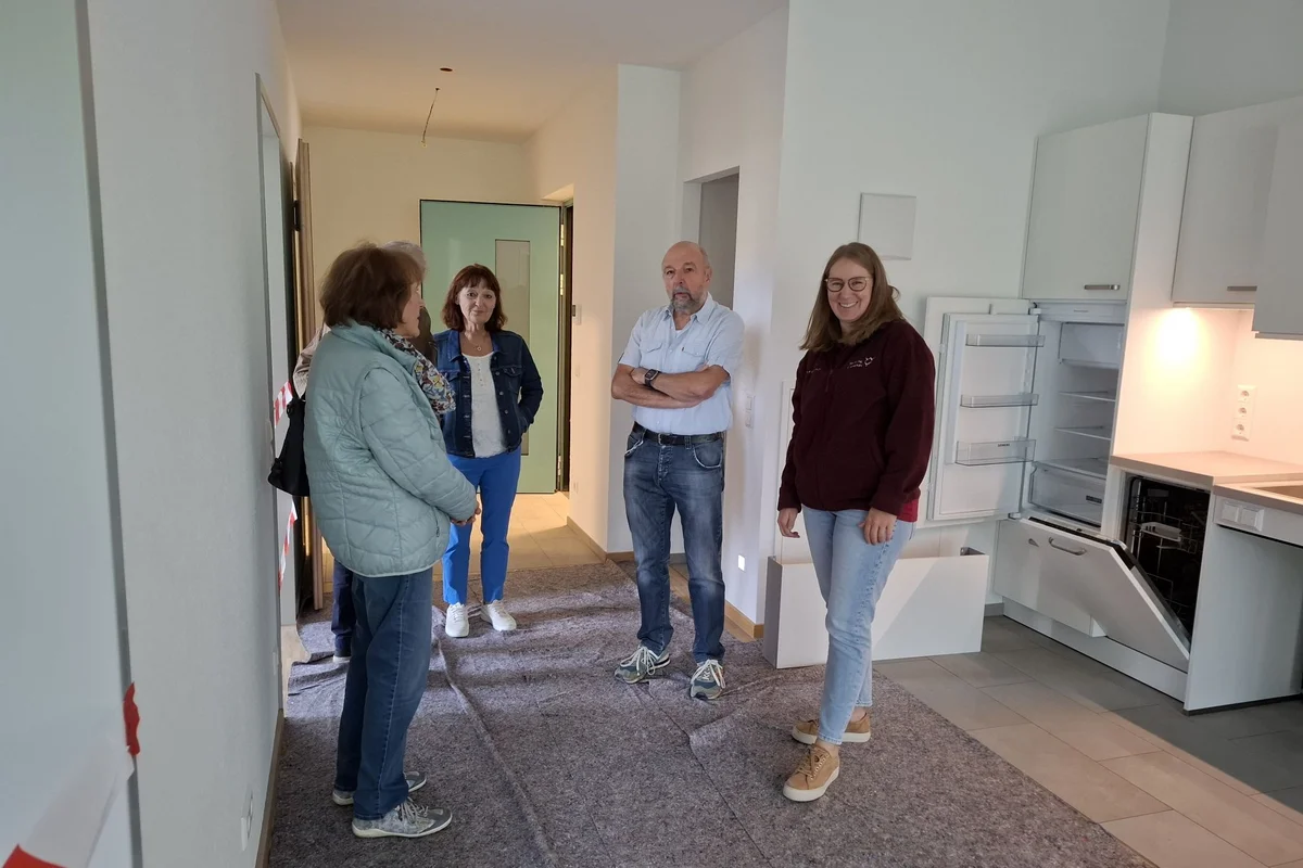 Tag der offenen Baustelle Fünf Personen besichtigen eine helle, halb eingerichtete Wohnung mit offener Küche; sie stehen im Flur und unterhalten sich, während Geräte im Küchenbereich offen sind.