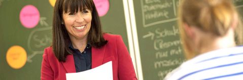 Das Bild zeigt eine Frau mit einem Paper vor einer Schultafel