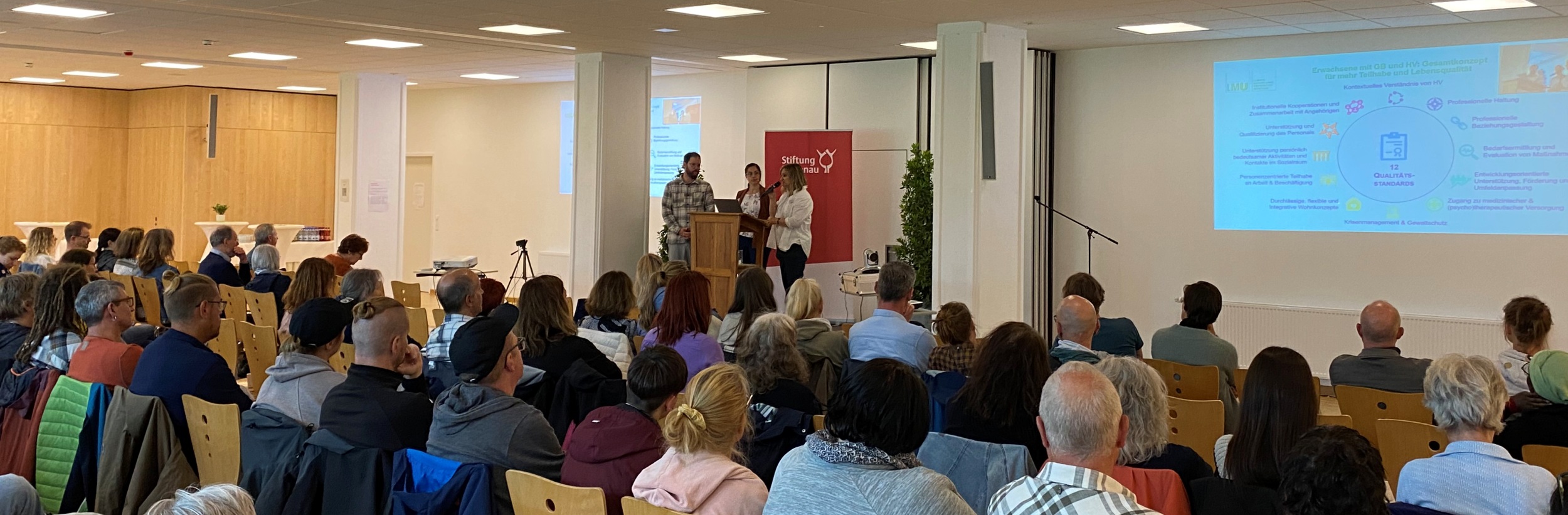 Das Bild zeigt mehrere Personen an einem Rednerpult, im Hintergrund eine Projektion, vor ihnen Zuhörer