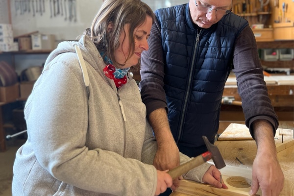 Besuch einer Zimmerei Das Bild zeigt eine Frau, die mit Hilfe eines Mannes einen Nagel in ein Brett schlägt