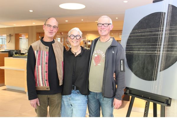 Gruppenfoto Auf dem Bild sieht man zwei Männer und eine Frau mi Brille, die zusammen in die Kamera schauen und lächeln. Neben ihnen steht ein Gemälde, auf dem ein Kreis zu sehen ist