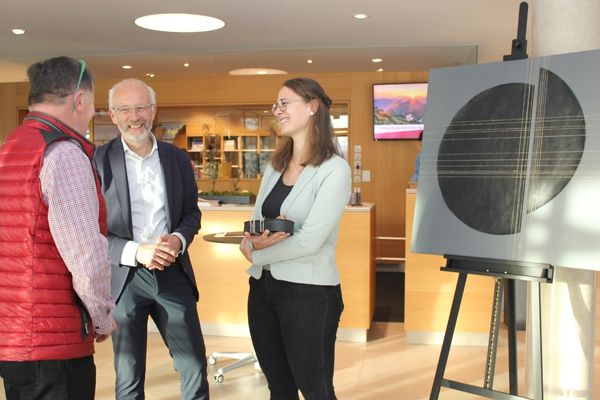 BM Smigoc Zwei Männer und eine Frau Stehen vor einem Ausstellungsstück, das eine Art Kreis zeigt und reden lächelnd miteinander.
