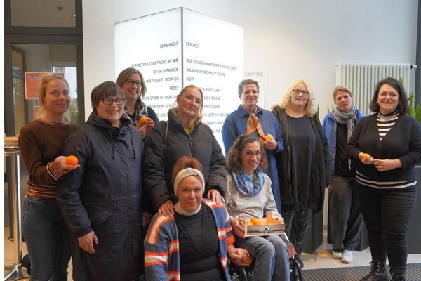 Lichtersäule Auf dem Bild sieht man viele Personen die alle versammelt vor einer Lichtersäule stehen. Sie halten Orangen in der Hand und lächeln in die Kamera