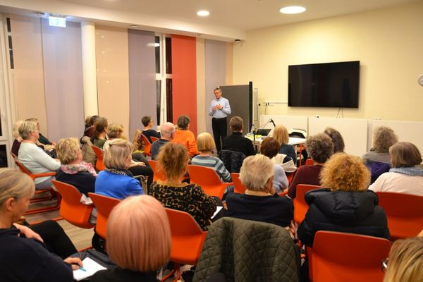 Vortrag Dr. Jochen Tenter Das Bild zeigt einen Raum in dem viele Personen sitzen auf orangenen Stühlen und einem Vortrag zuhören