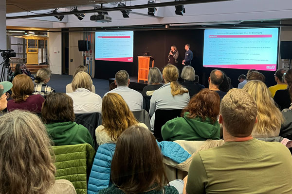 Zwei Personen tragen etwas vor und viele Personen hören zu.