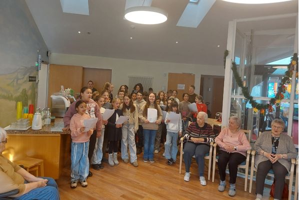Gemeinsam singen Das Bild zeigt eine Gruppe von Kindern die Lieder singen. Sie halten Liedzettel in der Hand um am Rand sitzen mehrere ältere Menschen und hören zu.