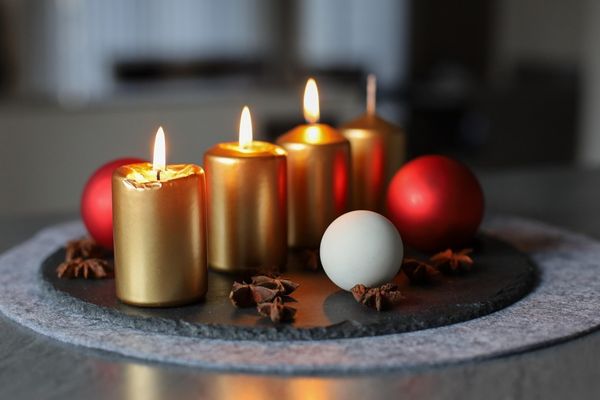 Das Bild zeigt vier goldene Kerzen die auf einem grauen Untersetzer stehen und brennen. Neben ihnen liegen Weihnachtskugeln in weiß und rot 