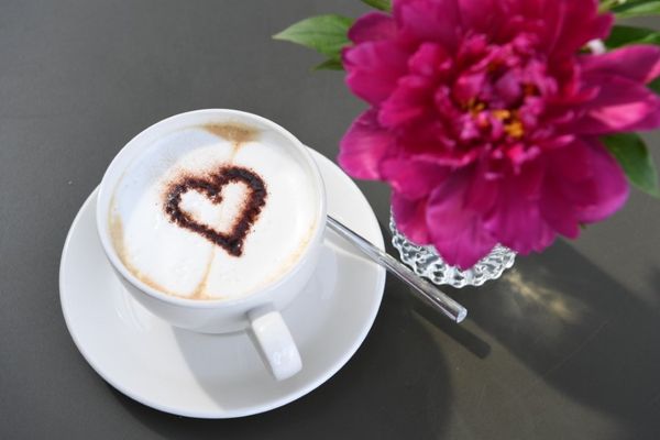 Auf dem Bild ist eine Kaffeetasse und eine pinke Blume zu sehen. Auf dem Schaum in der Tasse ist mit Kakaopulver ein Herz gezeichnet