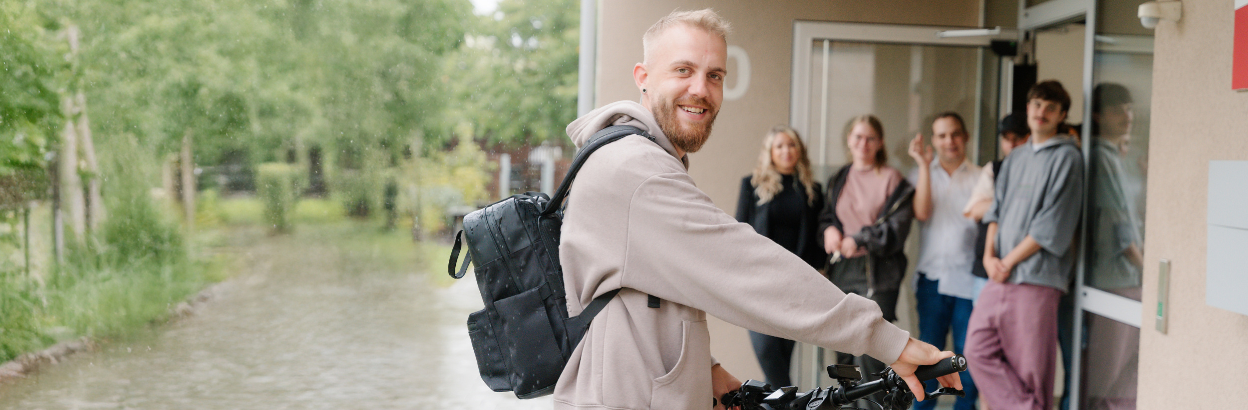 Das Bild zeigt einen Mann mit Rucksack vor einer Gruppe von Menschen an einem Hauseinang